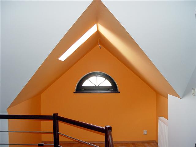 Pomarańczowa ściana na poddaszu z oknem w kształcie półkola, widok zza drewnianej barierki schodów.